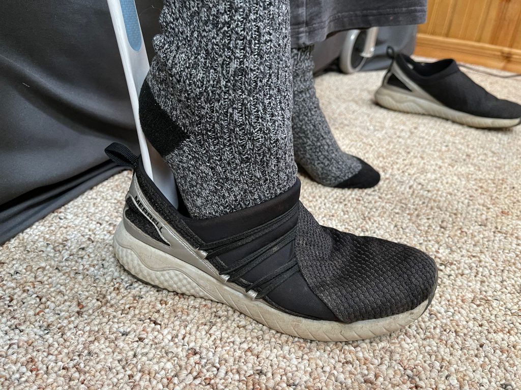 A close-up of a person using a long-handled shoehorn to assist in putting on a black slip-on shoe over a gray and black sock. The shoe features elastic straps for a snug fit, and the scene highlights the practicality of adaptive tools for individuals with limited mobility. Lower Body Dressing After Hip Replacement Surgery