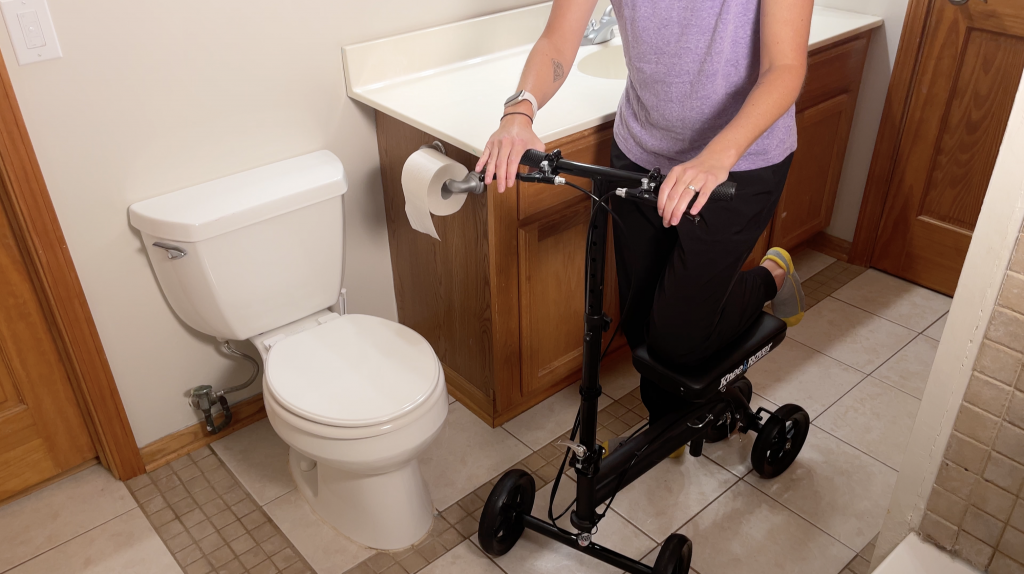 Person using a black knee scooter in a bathroom, positioning their non-injured leg on the inside near the toilet for better access. The scooter is designed for mobility while keeping the injured leg elevated and supported. How to Use a Knee Scooter in a Bathroom - EquipMeOT