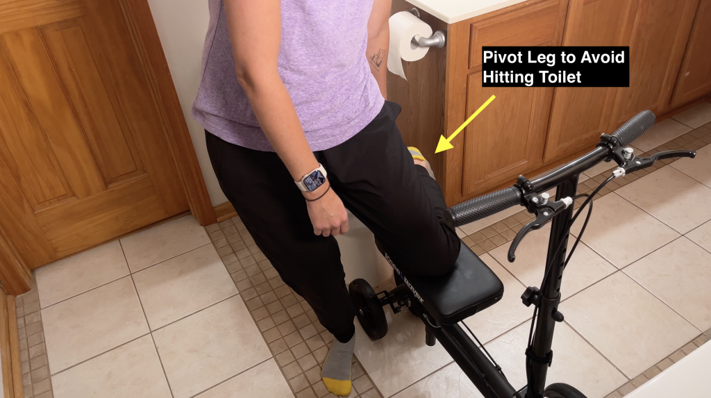 Person using a black knee scooter in a bathroom, pivoting their leg to avoid hitting the toilet. A yellow arrow highlights the action with the text 'Pivot Leg to Avoid Hitting Toilet' for instructional emphasis. How to Use a Knee Scooter in the Bathroom - EquipMeOT