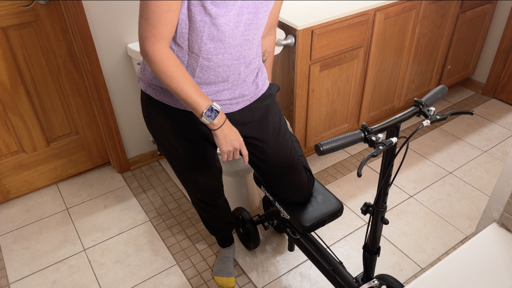 Person positioning a black knee scooter in a bathroom, backing it up toward the toilet. The scooter supports their injured leg while they prepare to sit safely. How to Use a Knee Scooter in the Bathroom - EquipMeOT