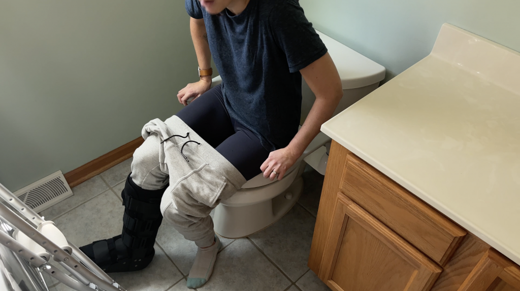 A person seated on a raised toilet seat equipped with safety rails, demonstrating safe usage for individuals with limited mobility or non-weight-bearing conditions. They are wearing a medical walking boot on one leg, with sweatpants pulled down and layered over black leggings. The individual is stabilizing themselves using the safety rails, showcasing an adaptive bathroom setup. How to Use Toilet When Non-Weight Bearing with Crutches or Walker - EquipMeOT