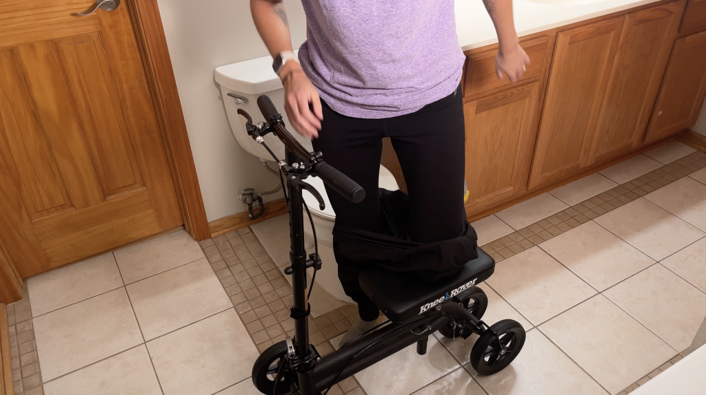 Person standing with the support of a black knee scooter in a bathroom, lowering their pants to knee height. The scooter provides stability while they prepare to use the toilet. How to Use Knee Scooter in the Bathroom - EquipMeOT