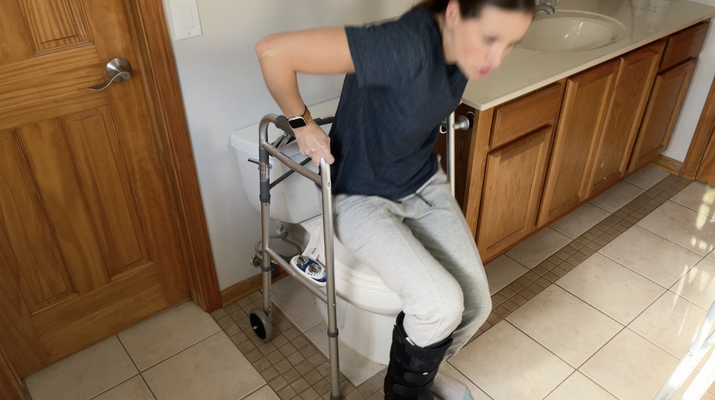 A person using a walker to push back and upward for standing up from the toilet while maintaining a non-weight-bearing position on the injured leg. How to Use Toilet When Non-Weight Bearing with Crutches or Walker - EquipMeOT