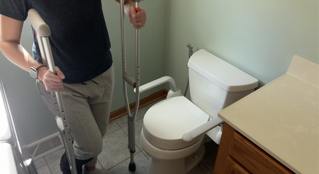 A person using crutches stands next to a toilet equipped with a raised toilet seat and safety rails in a bathroom. The person is wearing light gray sweatpants and a smartwatch, with natural light illuminating the space. How to Use Toilet When Non-Weight Bearing with Crutches or Walker - EquipMeOT