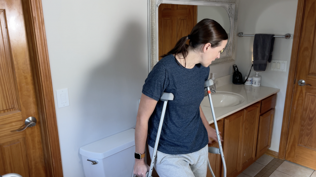 Person using crutches in a bathroom, positioning themselves near a toilet. The individual is wearing a dark blue shirt and light gray pants, preparing to use the counter behind them next to the toilet for balance. How to Use Toilet When Non-Weight Bearing with Crutches or Walker - EquipMeOt