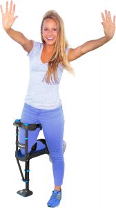 Smiling woman demonstrating the use of a hands-free knee crutch device. She is wearing a gray shirt, blue pants, and sneakers while balancing on the mobility aid, which supports her bent knee and allows for easy movement. How to use a knee scooter in the bathroom - EquipMeOT