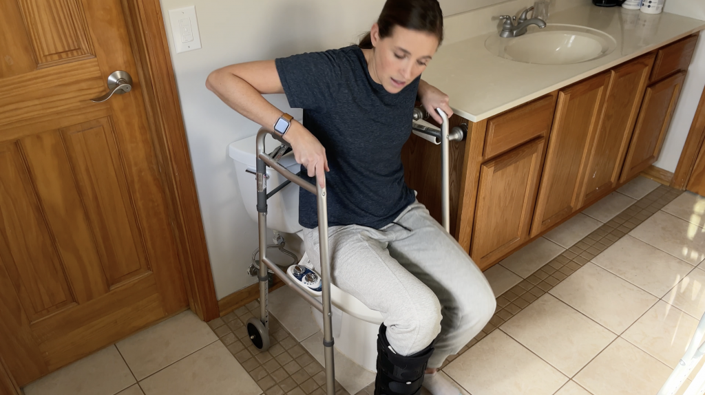 Person using walker handles for stability and leverage to push themselves up from the toilet while keeping their injured leg in a non-weight-bearing position. How to Use Toilet When Non-Weight Bearing with Crutches or Walker - EquipMeOT