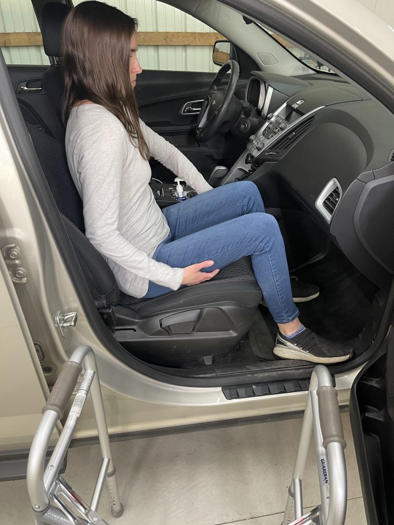 A woman sits in the passenger's seat of a car with a walker positioned outside the open door. She is grabbing her right leg with her right arm. She is dressed in a light gray long-sleeve shirt, blue jeans, and black slip-on shoes. The interior of the vehicle is visible, including the dashboard and steering wheel. The image illustrates a transition into or out of the car with the aid of mobility equipment. How to Get In and Out of the Car After Hip Replacement