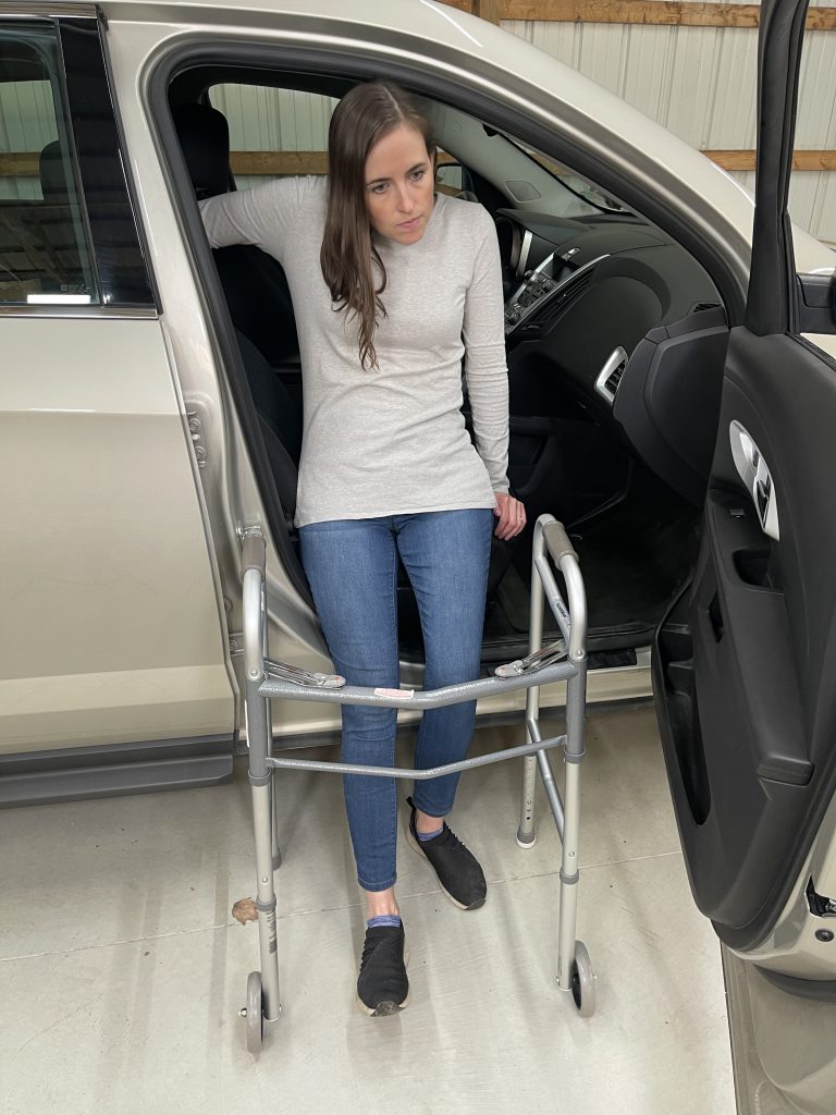 A woman carefully steps into a car using a walker for balance and support. She holds the car's door frame with one hand while steadying herself on the seat with the other. She is looking out and ducking her head underneath the car door frame. Dressed in a light gray long-sleeve shirt, blue jeans, and black slip-on shoes, she demonstrates a safe method for vehicle exit with mobility assistance. How to Get In and Out of the Car After Hip Replacement