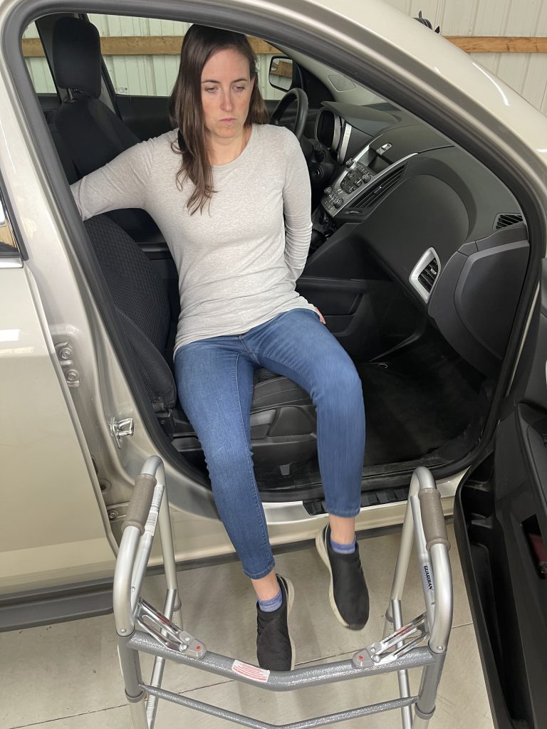 A woman steps out of a car with the aid of a walker. She uses one hand to hold the car seat for balance and the other to hold the car frame. She is dressed in a light gray long-sleeve shirt, blue jeans, and black slip-on shoes. The car door is open, revealing part of the vehicle's interior, and the walker is positioned for support during the transition. How to Get In and Out of the Car After Hip Replacement