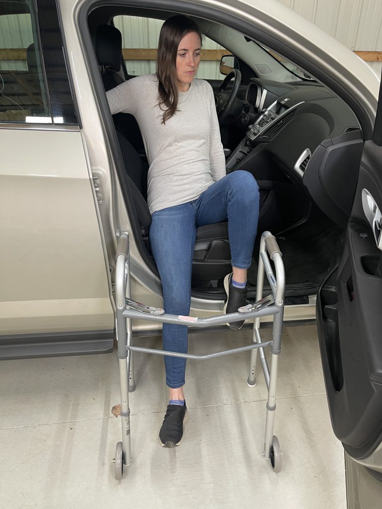 A woman is getting into a car, using a walker positioned in front of her for support. Her right leg is straight while her left leg is pushing off the running board. Dressed in a light gray long-sleeve shirt, blue jeans, and black slip-on shoes, she demonstrates a careful method for transitioning into a vehicle with mobility assistance. The car door is open, showing part of the interior. How to Get In and Out of the Car After Hip Replacement