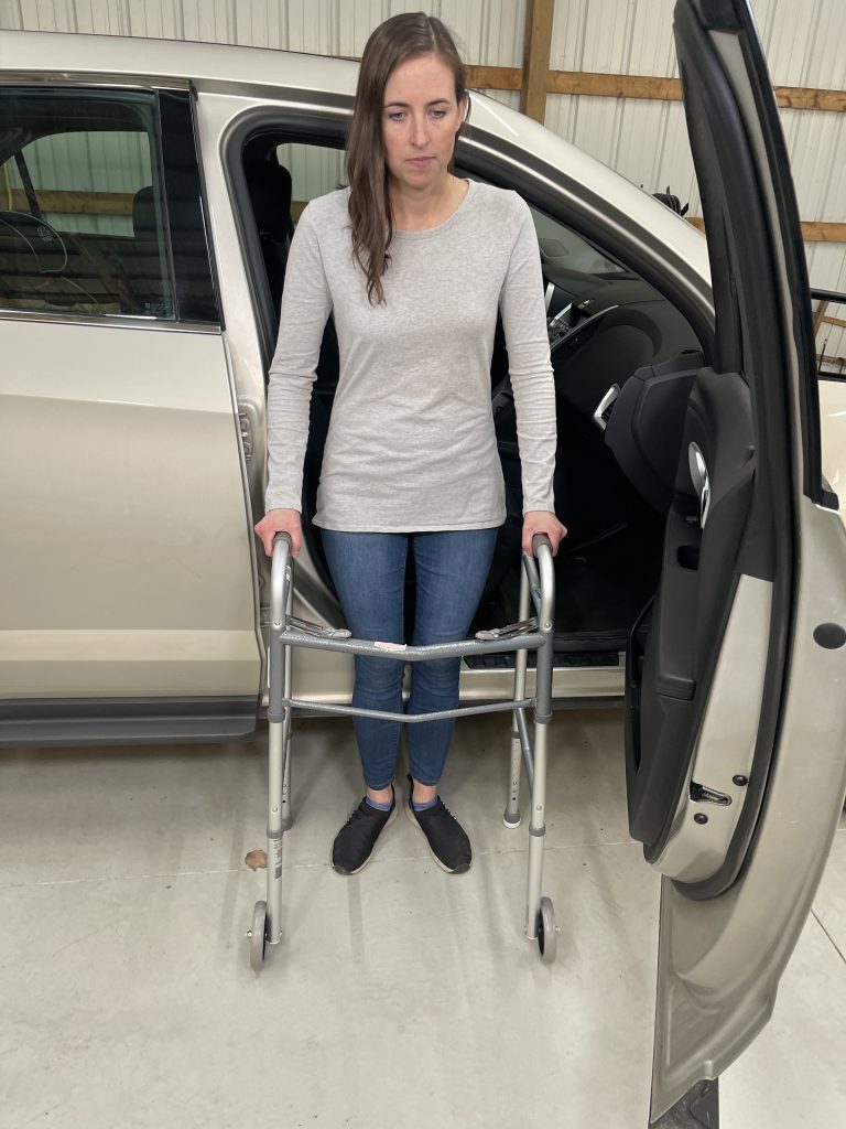 A woman stands backed up to the passenger seat outside a car holding onto a walker for support. She is wearing a light gray long-sleeve shirt, blue jeans, and black slip-on shoes. The car door is open, revealing part of the vehicle's interior. She is facing away from the car. How to Get In and Out of the Car After Hip Replacement