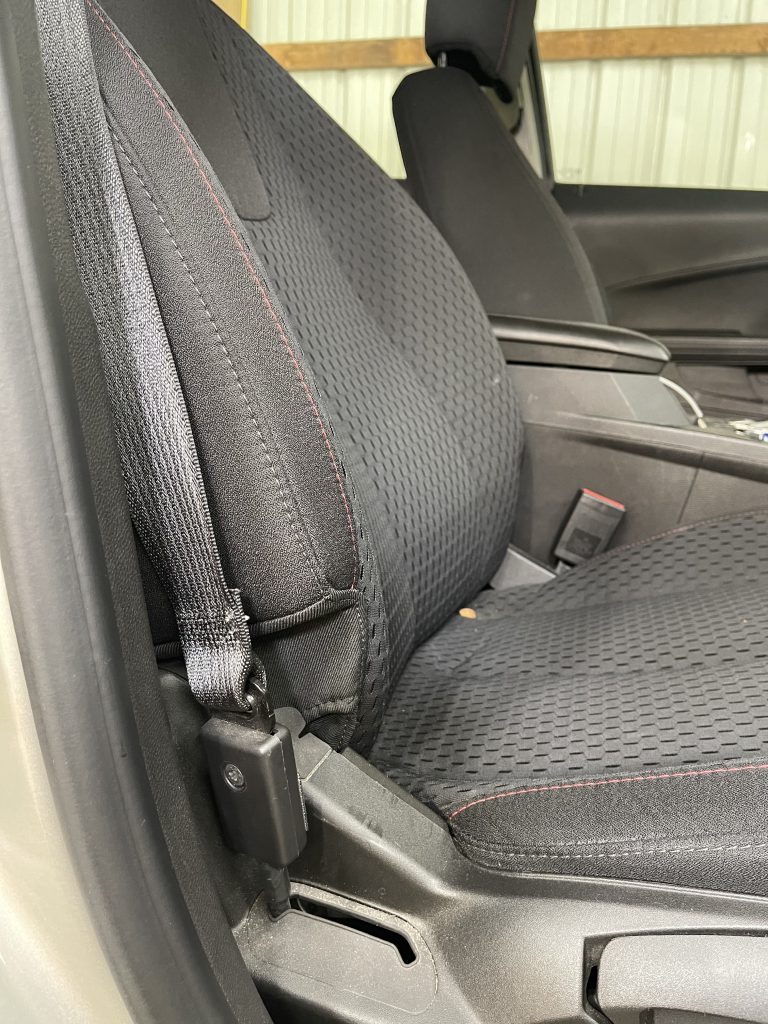 A close-up of a car seat with a black seatbelt extension device attached to the seatbelt mechanism. The seat features a textured black fabric with red stitching, and part of the car interior is visible, including the armrest and side panel. This setup highlights an adaptive seatbelt solution for improved accessibility or ease of use. How to Get In and Out of the Car After Hip Replacement